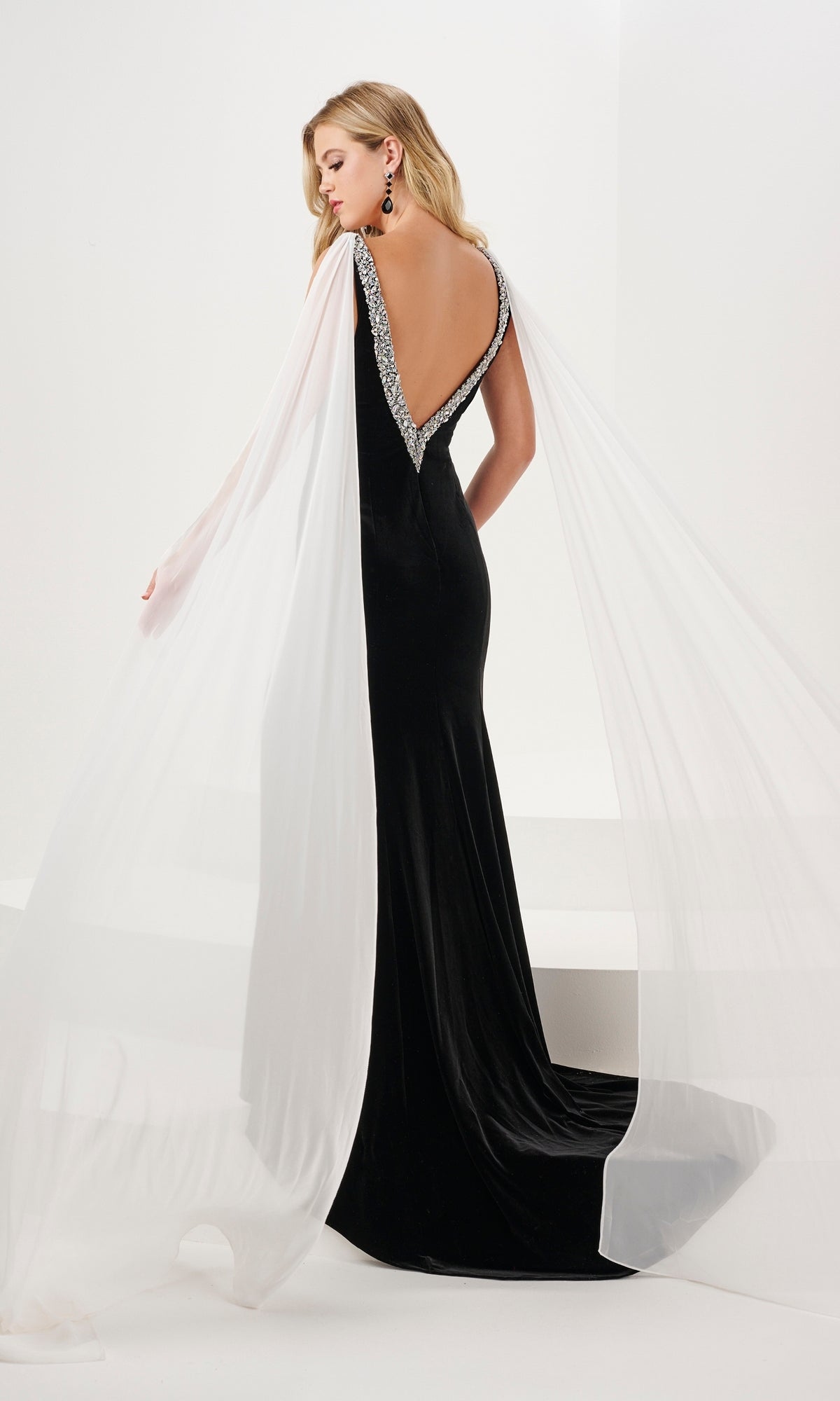 A woman wears the Long Prom Dress 14198 by Panoply, featuring stretch velvet, an open back, deep V neckline with silver trim, and sheer white cape-like fabric draped from the shoulders.